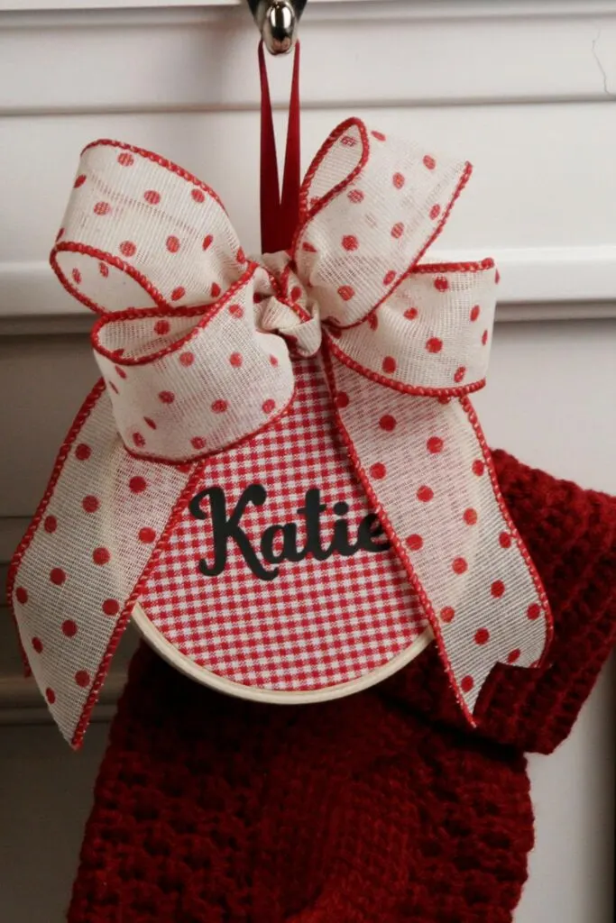 Embroidery hoop with red and white check fabric with the name Kate on it and a bow attached to a stocking