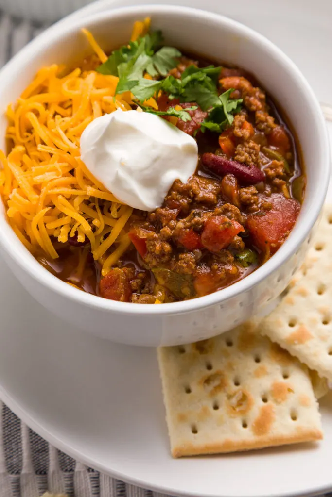 A bowl of chili topped with sour cream, shredded cheddar cheese and cilantro served with saltine crackers 