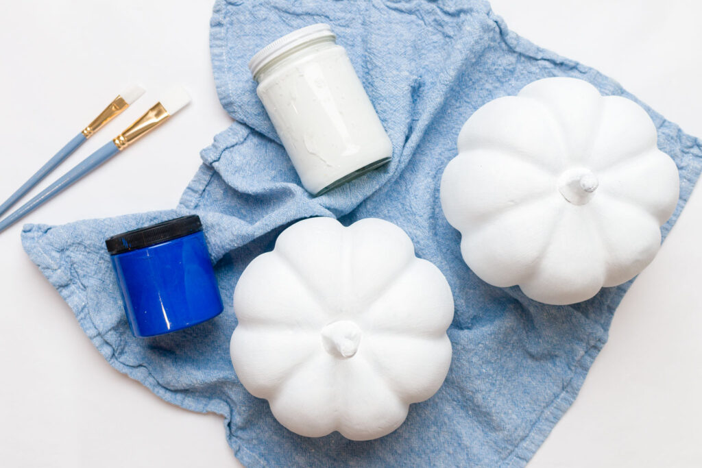 Two white pumpkins, white and blue paints, and paintbrushes on a table 