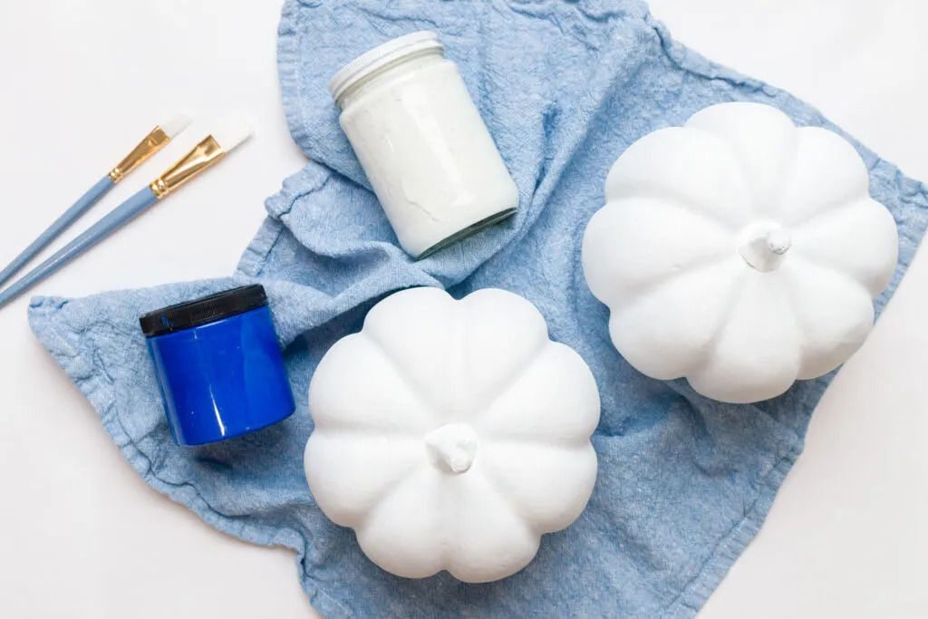 Two white pumpkins, white and blue paints, and paintbrushes on a table 