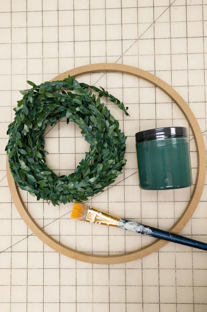 Wreath form, green paint and paintbrush on a craft mat 