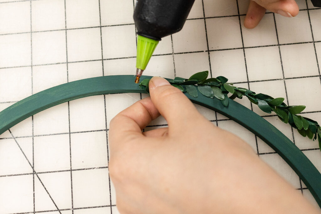 Using hot glue to attach the greenery to the painted wreath form