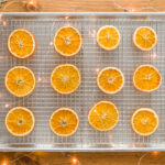 Dried Orange Slices on a wire rack in a sheet pan sitting on a wood table with fairy lights and snowflake oraments
