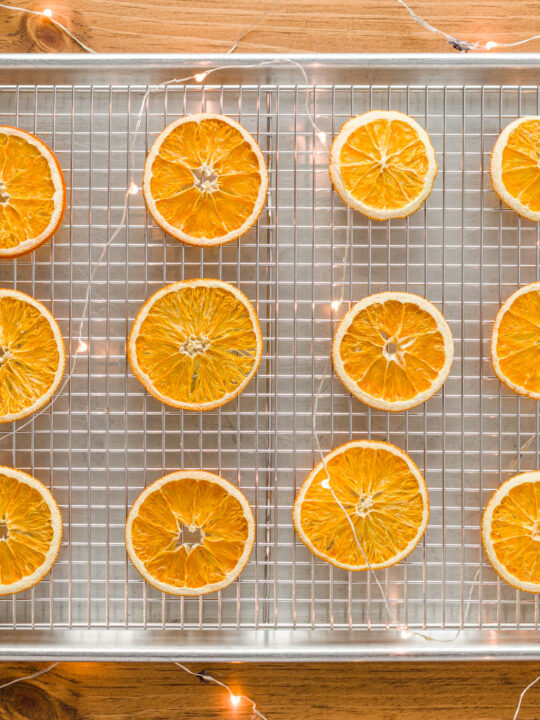 Dried Orange Slices on a wire rack in a sheet pan sitting on a wood table with fairy lights and snowflake oraments