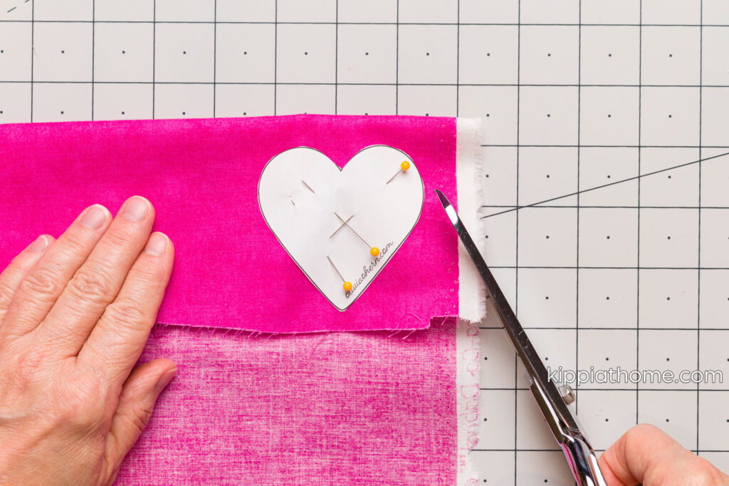 Cutting out the pinned heart pattern out of bring pink fabric with scissors on a cutting mat
