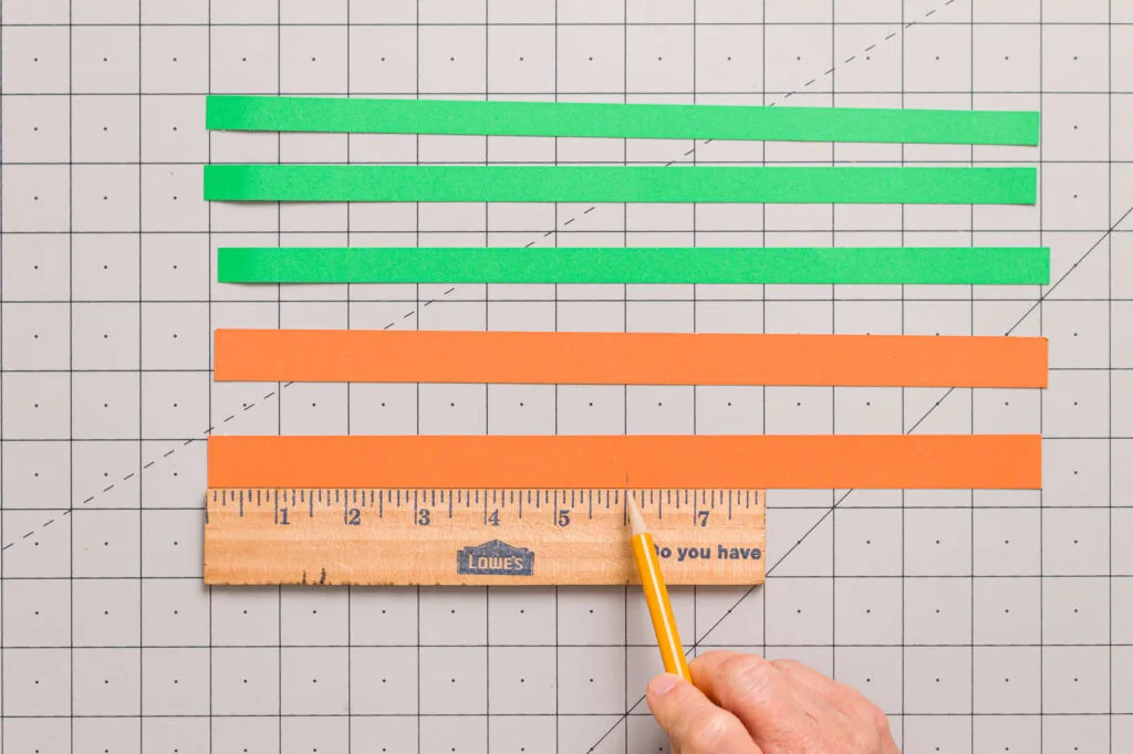 Marking the cut lines with a pencil on Green and orange paper strips lined up on a craft mat. 