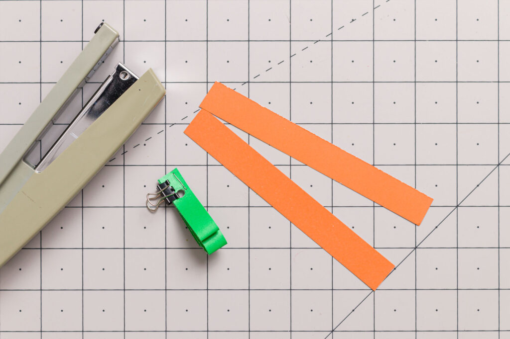 Orange paper strips and green folded paper strips held together with staple and clip on a craft mat