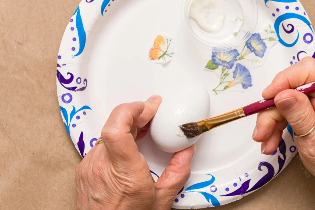 Using a paintbrush to apply Mod Podge to the egg on a paper plate