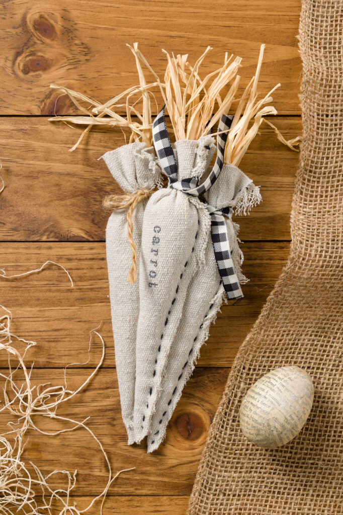three fabric Carrots placed in a bundle with straw , burlap, and an egg on a wooden table 