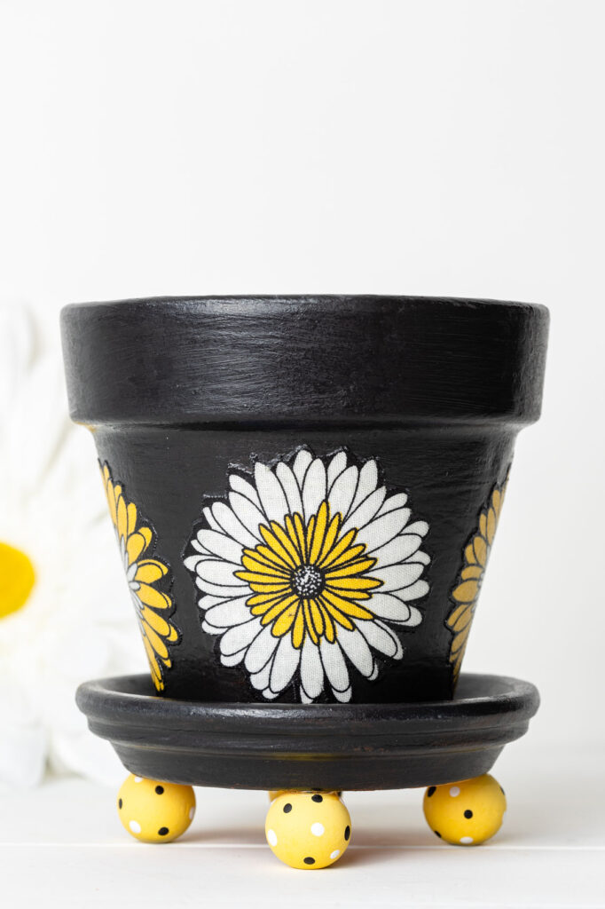 painted flower pot with fabric flowers added. Sitting on a painted saucer with wood bead feet