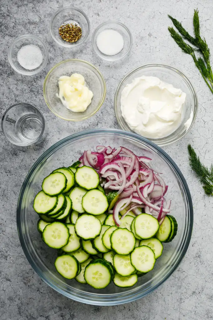 Sliced cucumbers and onions in a bowl with dressing ingredients in small bowls